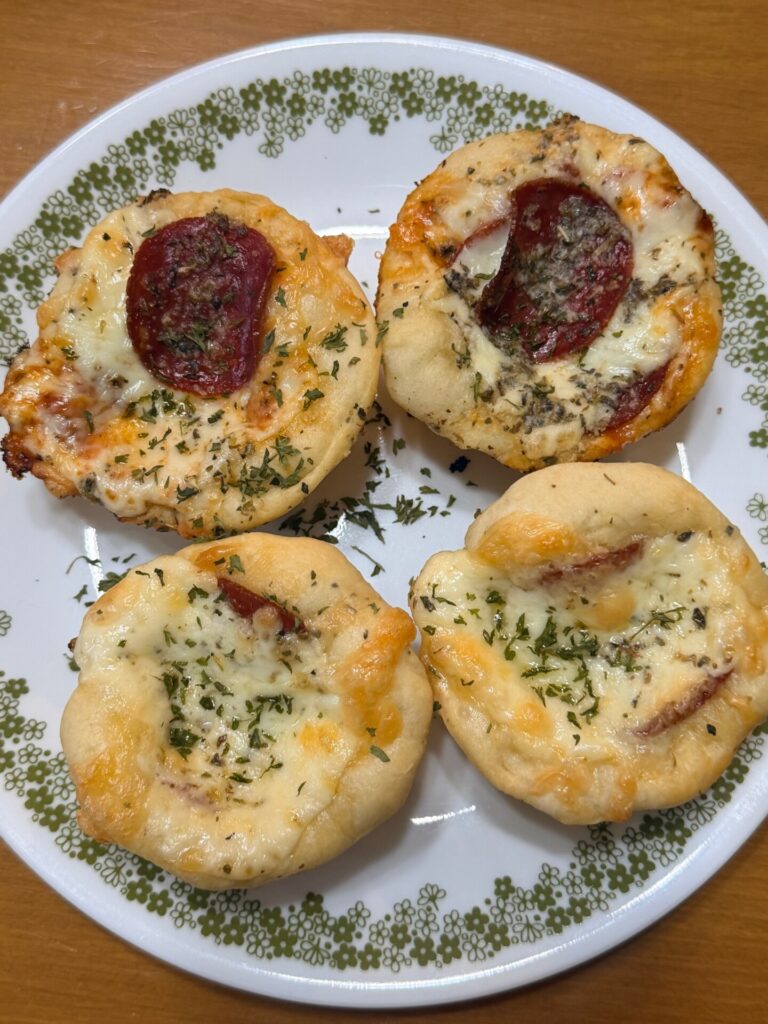 A white plate with a decorative green border holds four homemade mini pizzas topped with melted cheese, herbs, and slices of pepperoni on a wooden table.