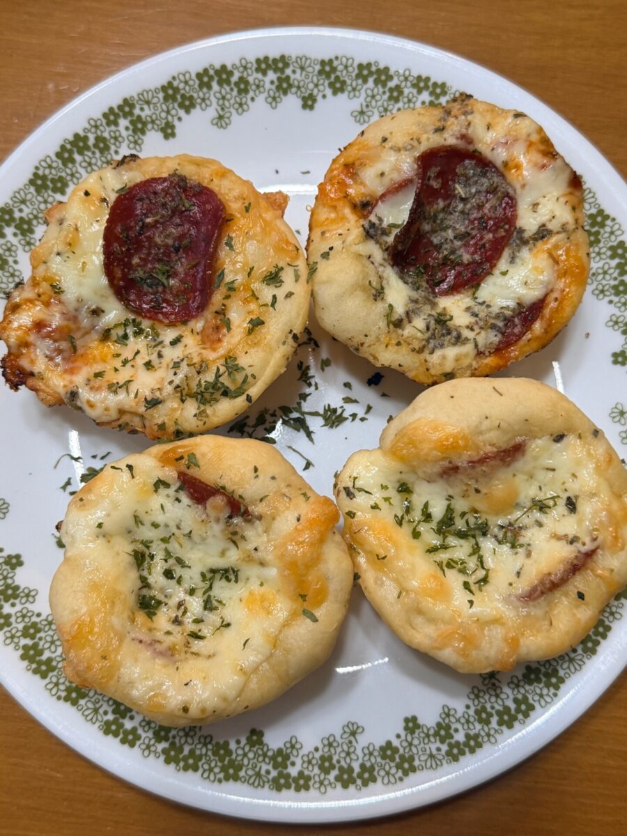 A white plate with a decorative green border holds four homemade mini pizzas topped with melted cheese, herbs, and slices of pepperoni on a wooden table.