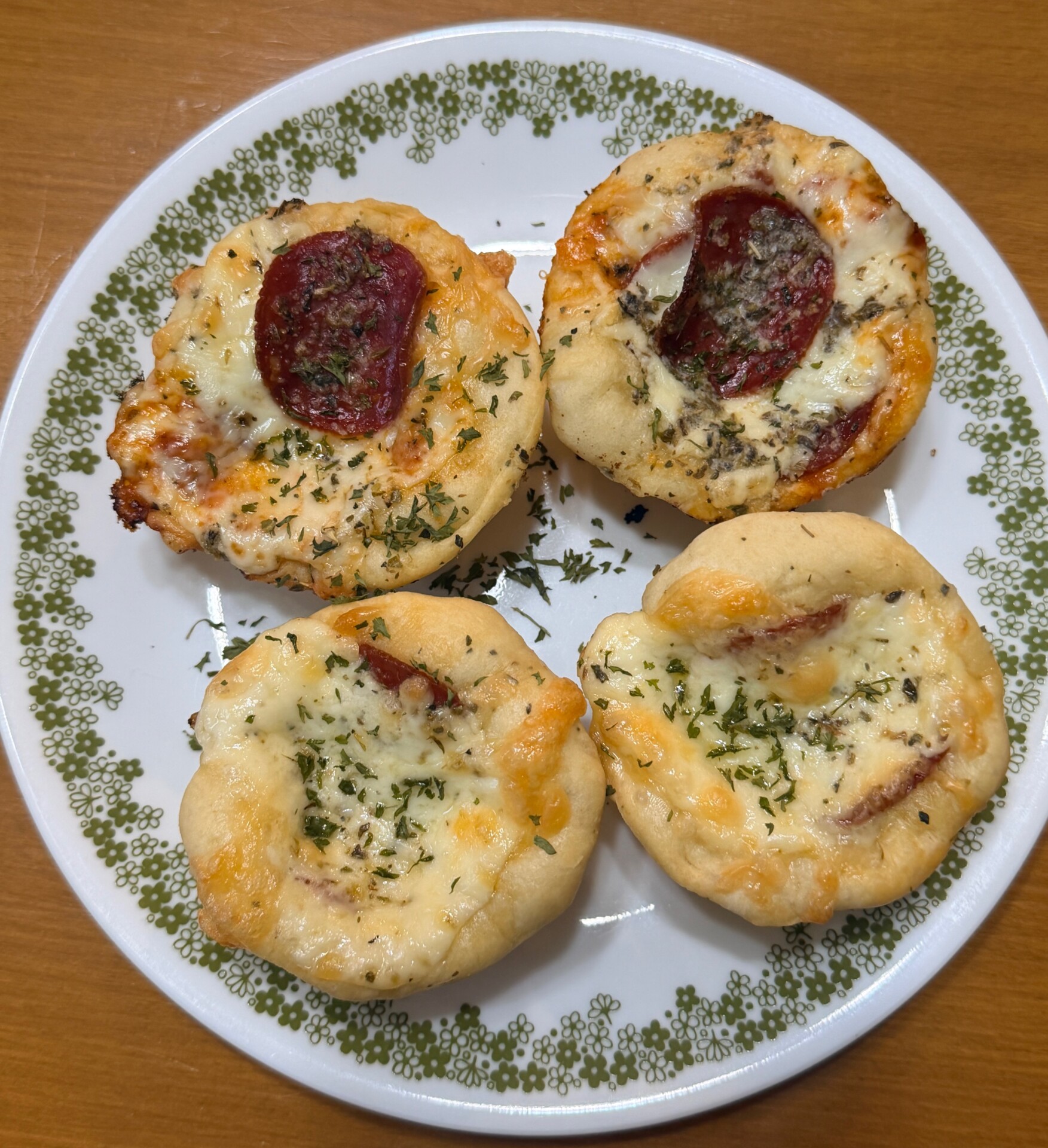 A white plate with a green floral border holds four small, round mini pizzas topped with melted cheese, herbs, and slices of pepperoni, placed on a wooden surface.