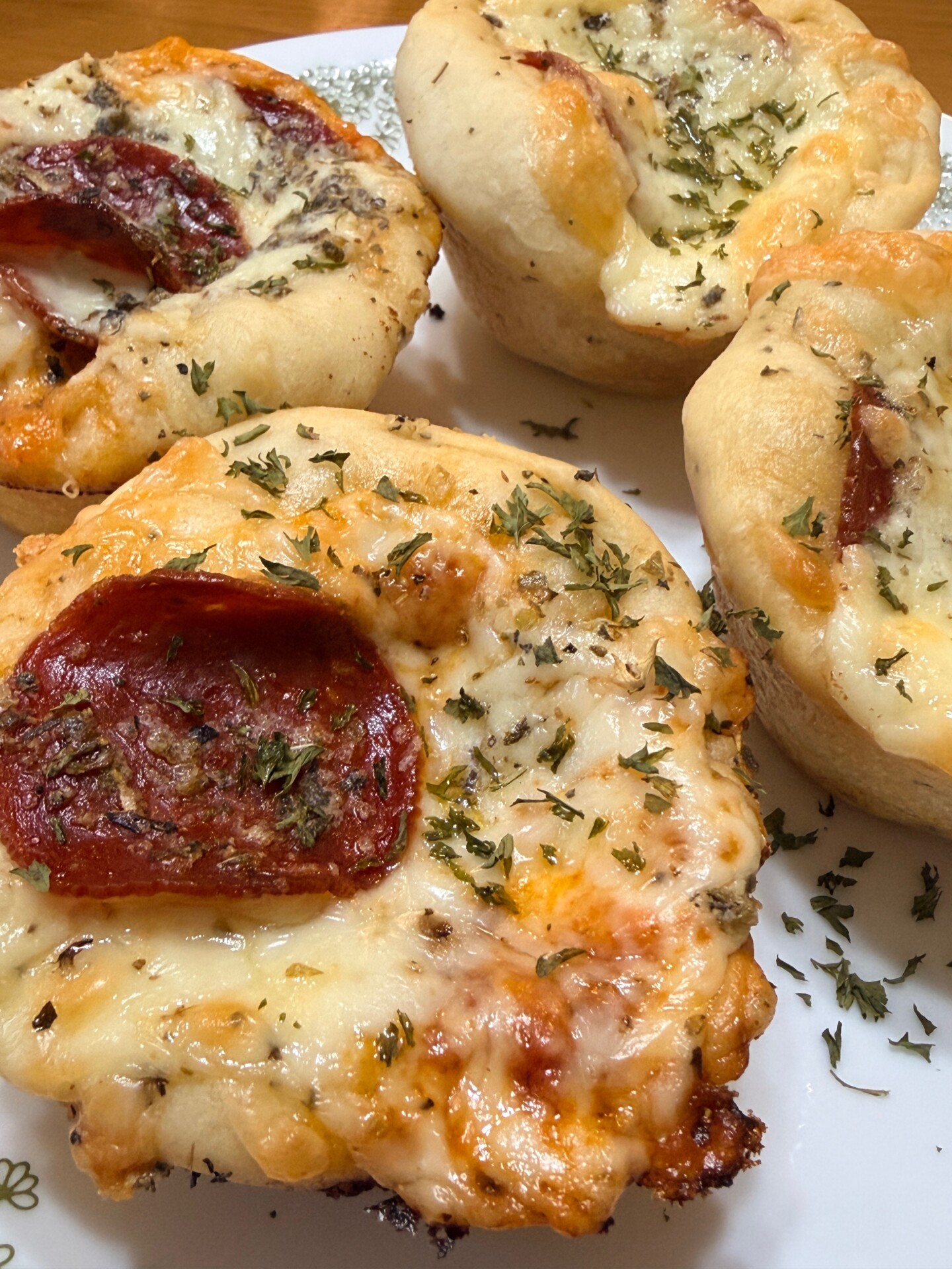 Four small, round, homemade pizzas topped with melted cheese, pepperoni, herbs, and baked in individual portions are arranged on a white plate with a decorative green border.