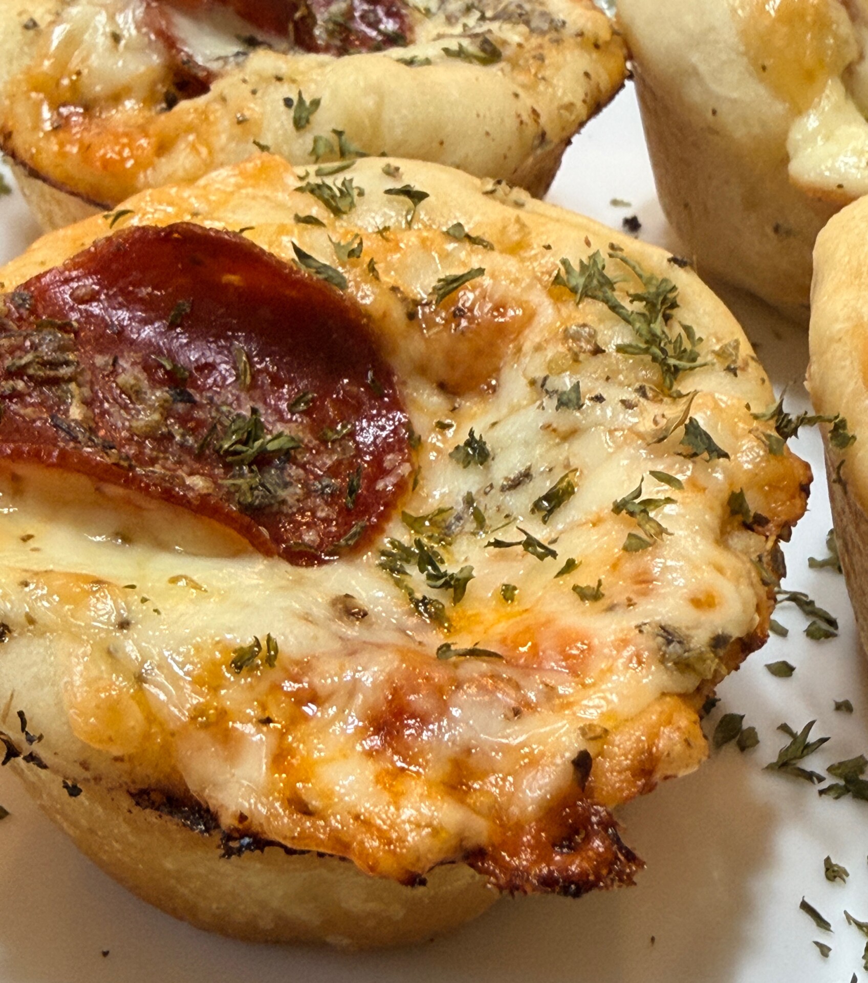 Close-up of mini pizza muffins topped with melted cheese, pepperoni, and sprinkled herbs, baked in golden-brown dough.