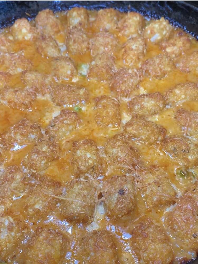 Golden brown tater tots are baked in a creamy, orange-hued cheese sauce, filling a skillet or baking dish. The melted cheese bubbles around the crispy tots, creating an inviting comfort food appearance.