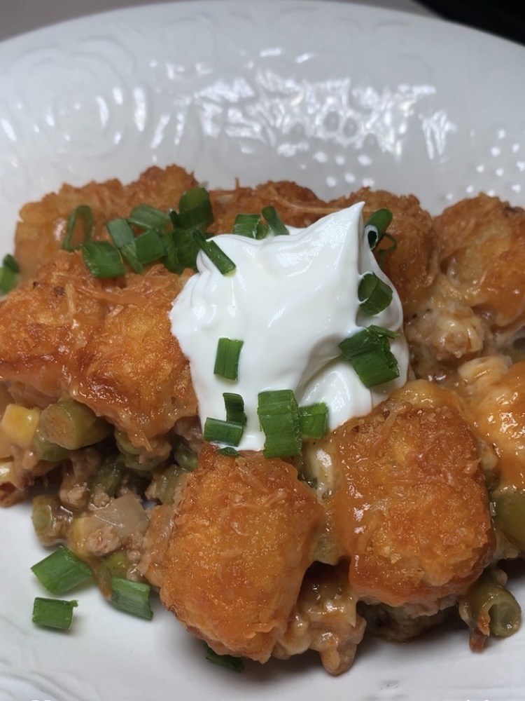 A close-up of tater tot casserole topped with a dollop of sour cream and chopped green onions, served on a white plate. The casserole includes golden tater tots, ground meat, cheese, and vegetables.