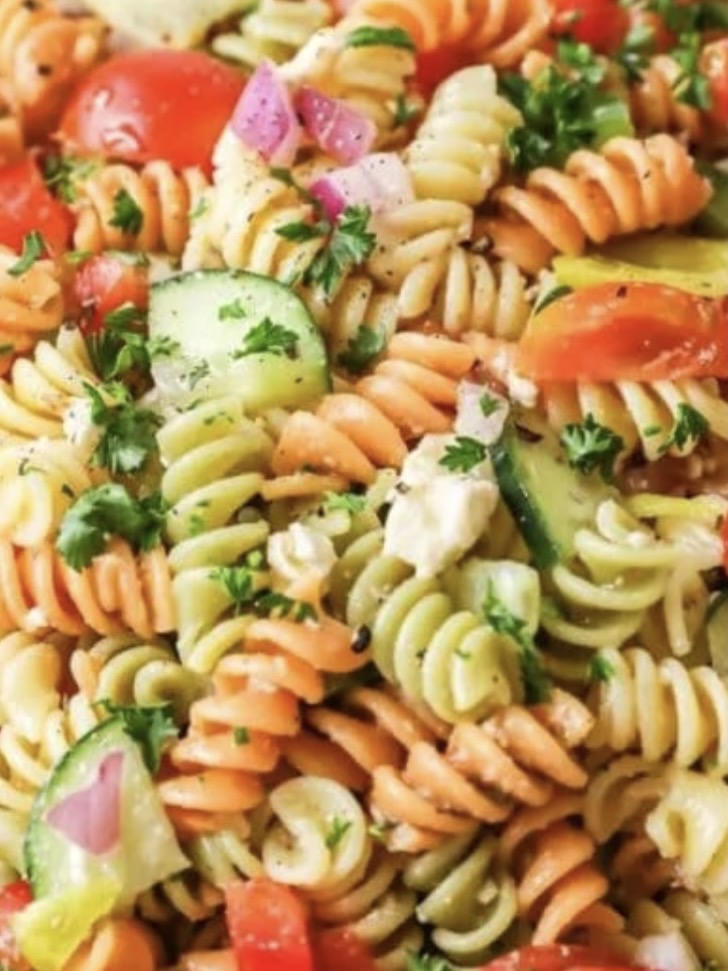 A close-up of colorful rotini pasta salad mixed with chopped cucumbers, tomatoes, red onions, parsley, and feta cheese. The salad is seasoned with black pepper and looks fresh and vibrant.
