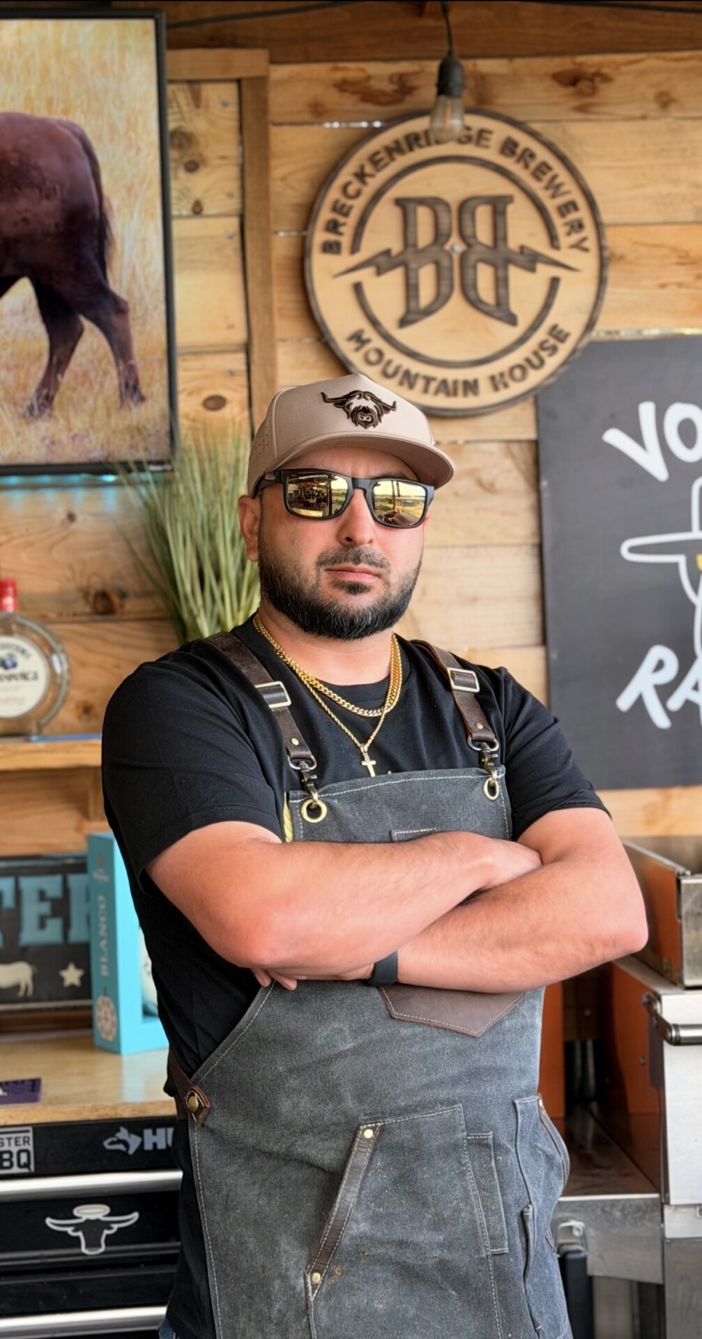 A man wearing sunglasses, a cap, and a dark apron stands with arms crossed in front of a wooden wall with a Breckenridge Brewery Mountain House sign. Bottles and a TV with a cow image are visible in the background.