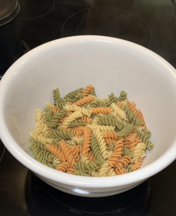 A white bowl filled with uncooked tri-color rotini pasta sits on a black stovetop. The pasta is a mix of green, orange, and yellow spirals.