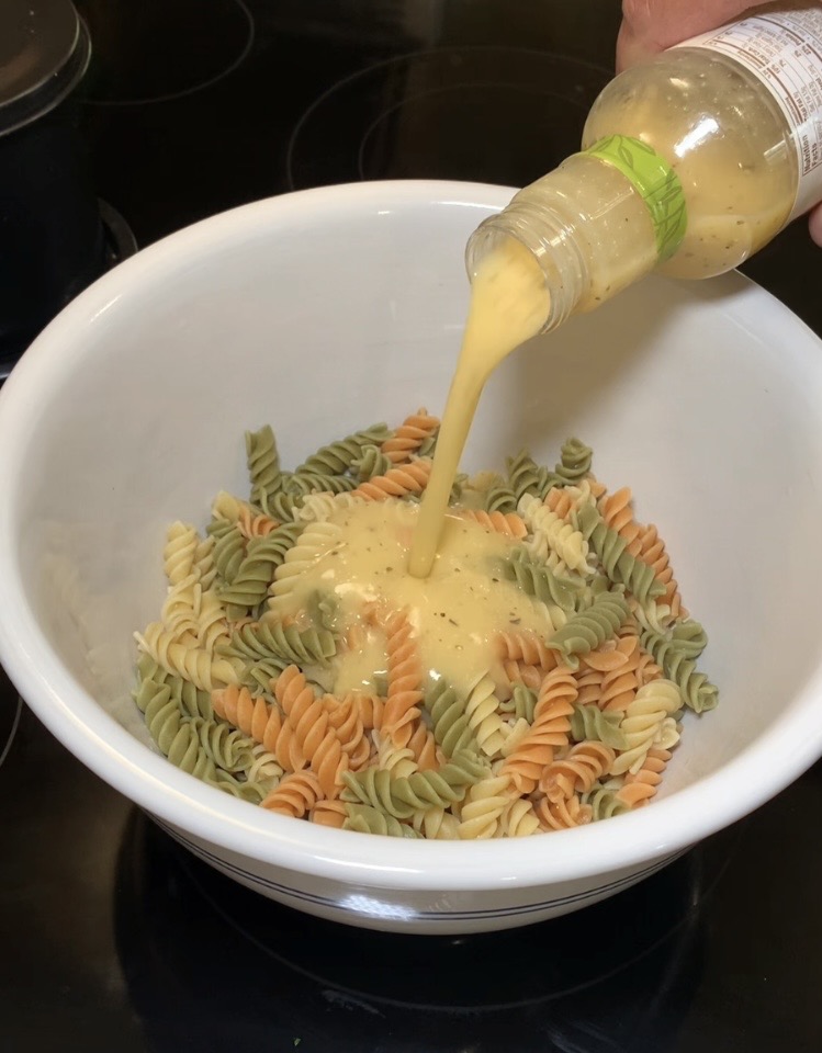 A hand pours creamy yellow dressing from a bottle onto a bowl of uncooked tri-color rotini pasta, which includes orange, green, and white spirals, in a white mixing bowl on a dark countertop.