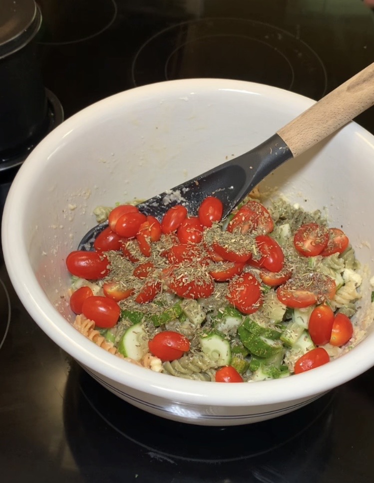 A white bowl on a stovetop contains pasta salad with cherry tomatoes, cucumber slices, spinach, and herbs, being mixed with a black spatula.