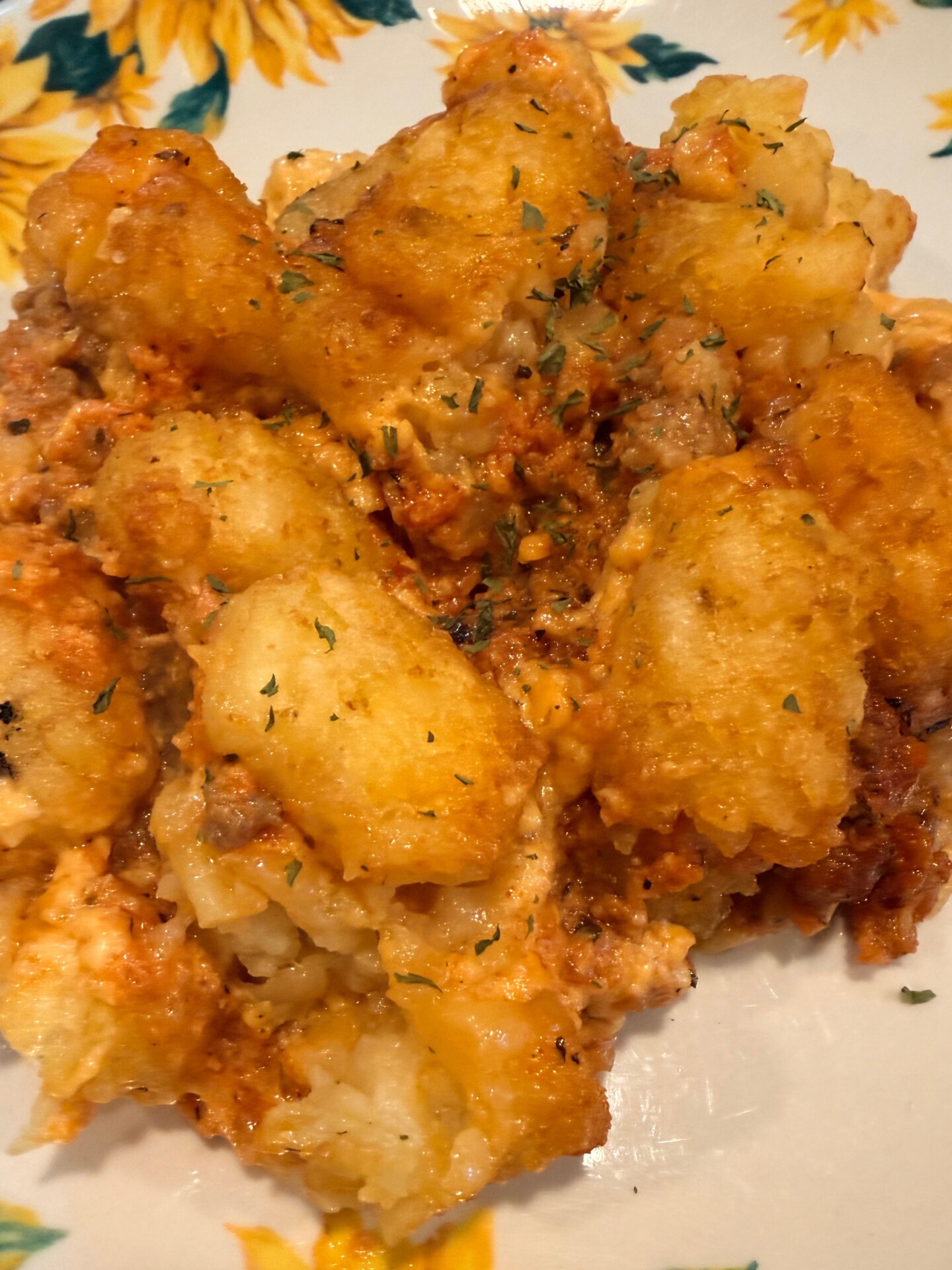 A close-up of a serving of tater tot casserole with golden-brown tater tots, melted cheese, ground meat, and herbs, on a floral-patterned plate.
