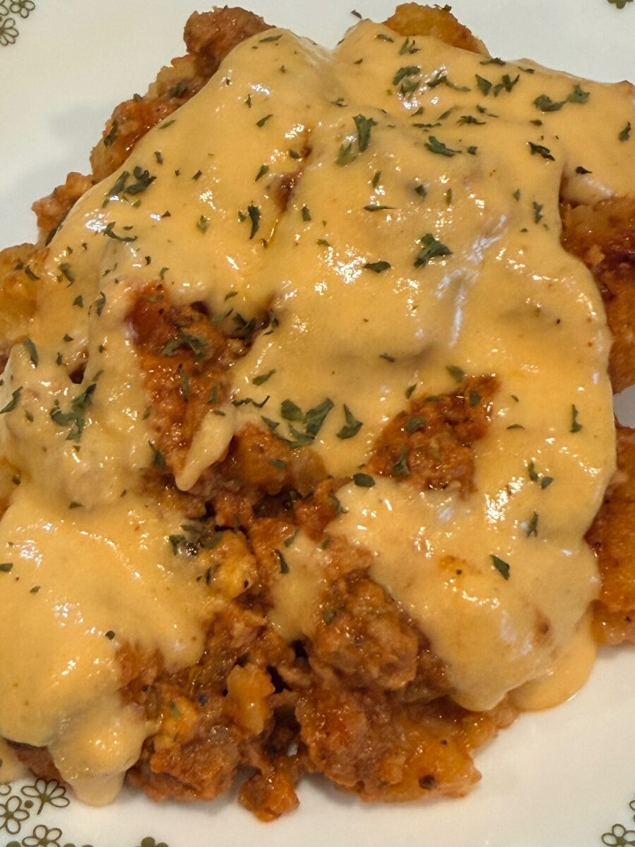A close-up of a savory dish topped with creamy orange cheese sauce, ground meat, and garnished with chopped herbs, served on a white plate with a subtle floral pattern.