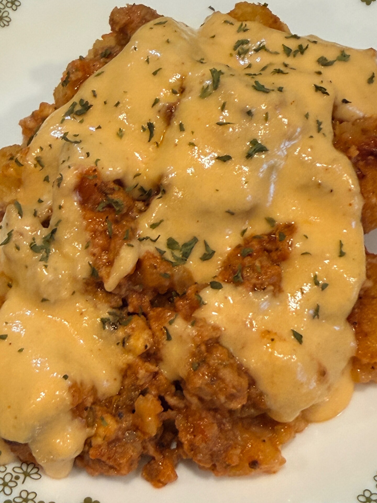 A close-up of a savory dish topped with creamy orange cheese sauce, ground meat, and garnished with chopped herbs, served on a white plate with a subtle floral pattern.