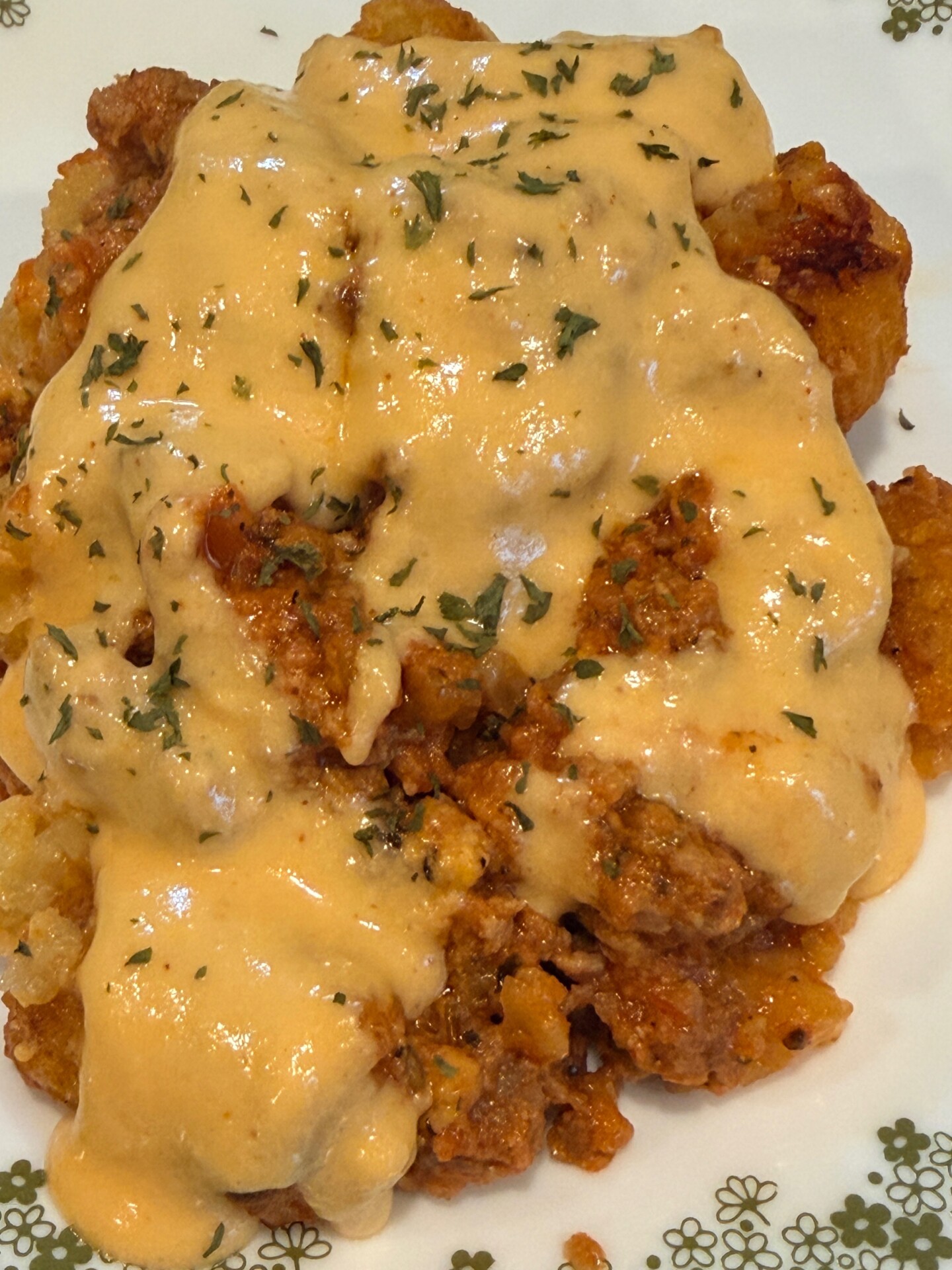 A close-up of a plate of cheesy tater tot casserole topped with ground meat sauce and sprinkled with chopped herbs. The melted cheese sauce covers most of the casserole, giving it a creamy appearance.