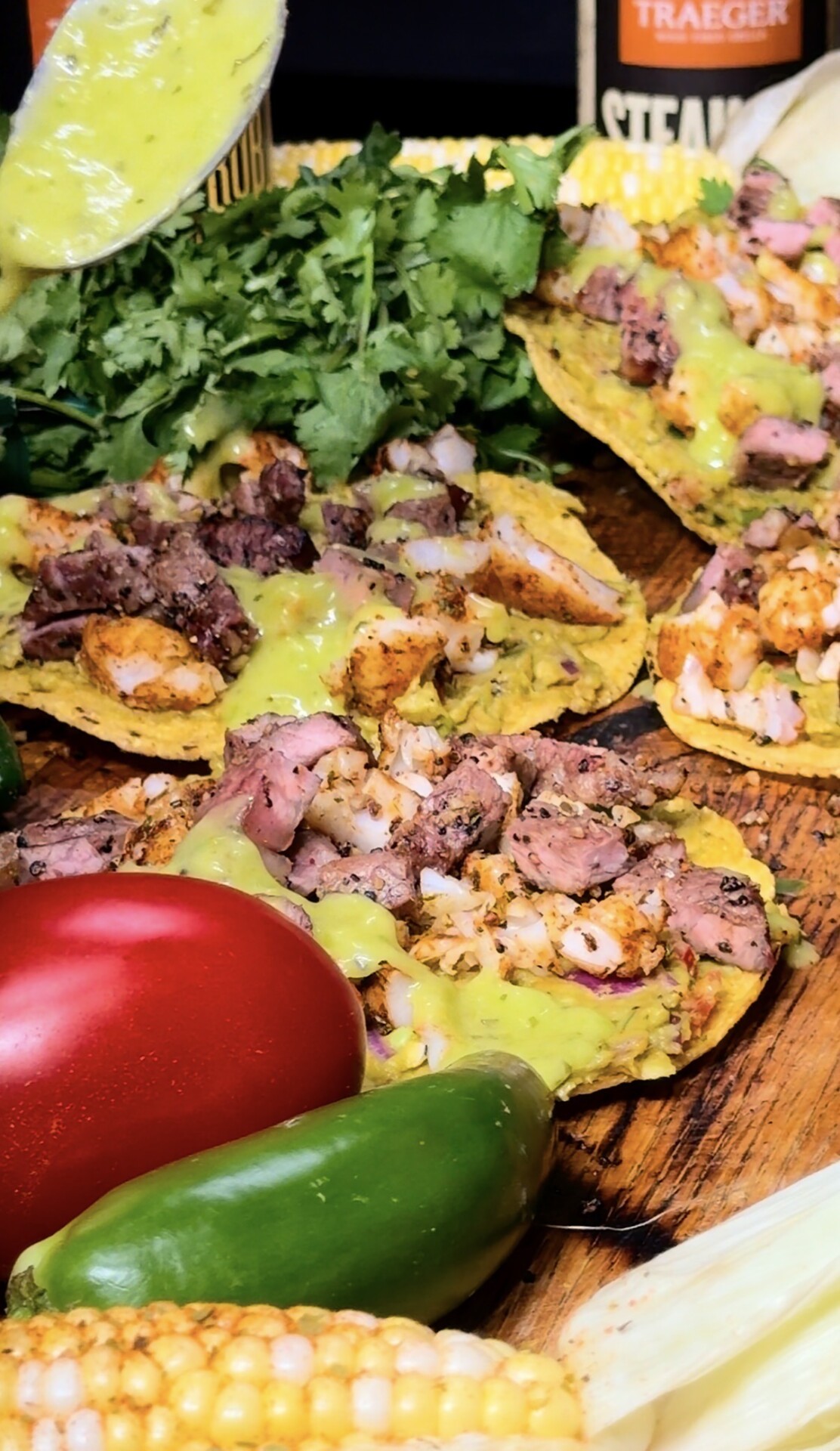 Three tostadas topped with grilled meat, green sauce, and chopped onions on a wooden surface, surrounded by fresh cilantro, a red bell pepper, jalapeños, corn, and a spoon pouring sauce.