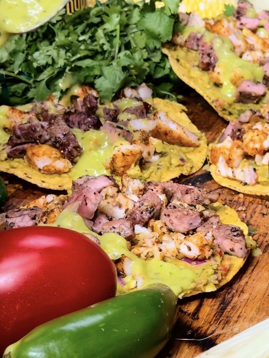 Tostadas topped with grilled meat, diced onions, and green sauce sit on a wooden surface, surrounded by fresh cilantro, a red tomato, and a green jalapeño pepper.
