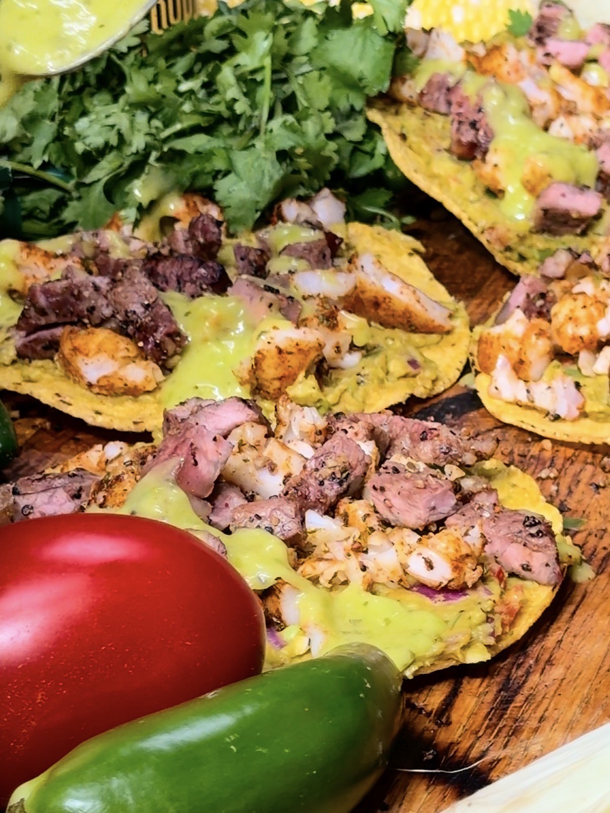 Tostadas topped with grilled meat, diced onions, and green sauce sit on a wooden surface, surrounded by fresh cilantro, a red tomato, and a green jalapeño pepper.