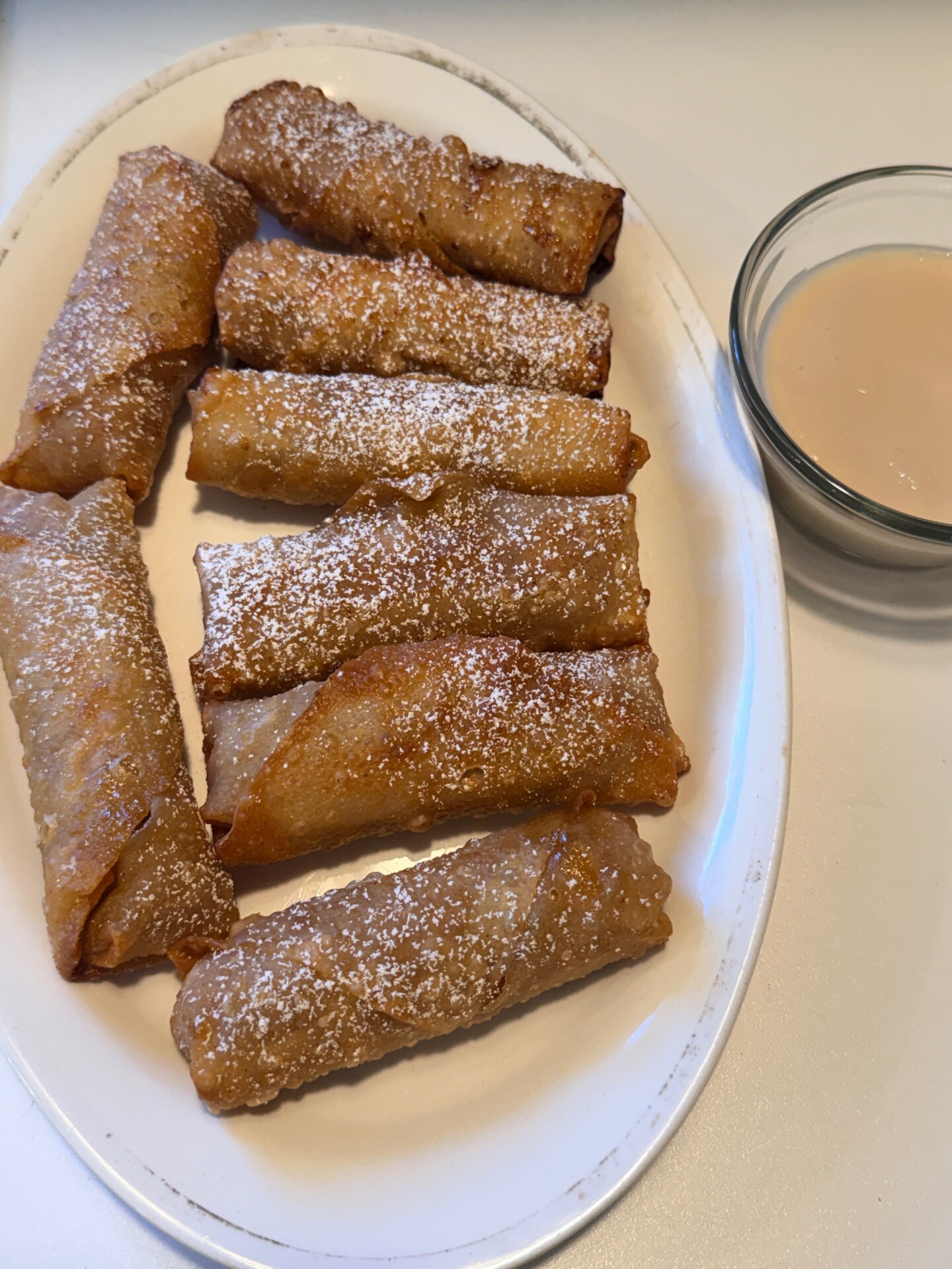 A white oval plate holds eight golden-brown, powdered sugar-dusted fried rolls, with a small glass bowl of creamy dipping sauce on the side.