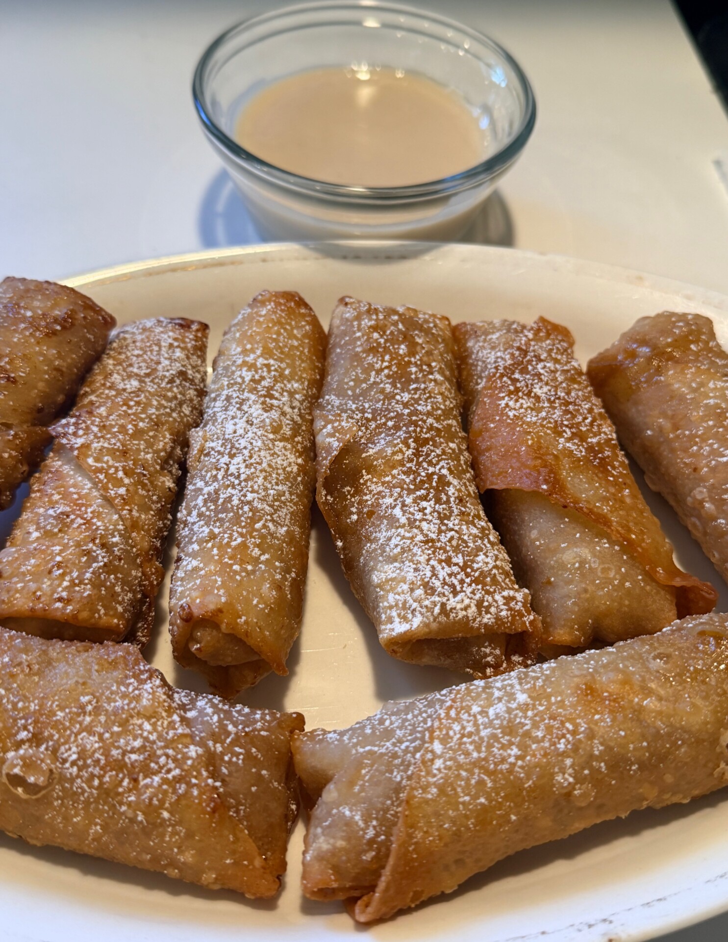 A plate of golden, crispy rolled pastries dusted with powdered sugar, served with a small glass bowl of light-colored dipping sauce in the background.