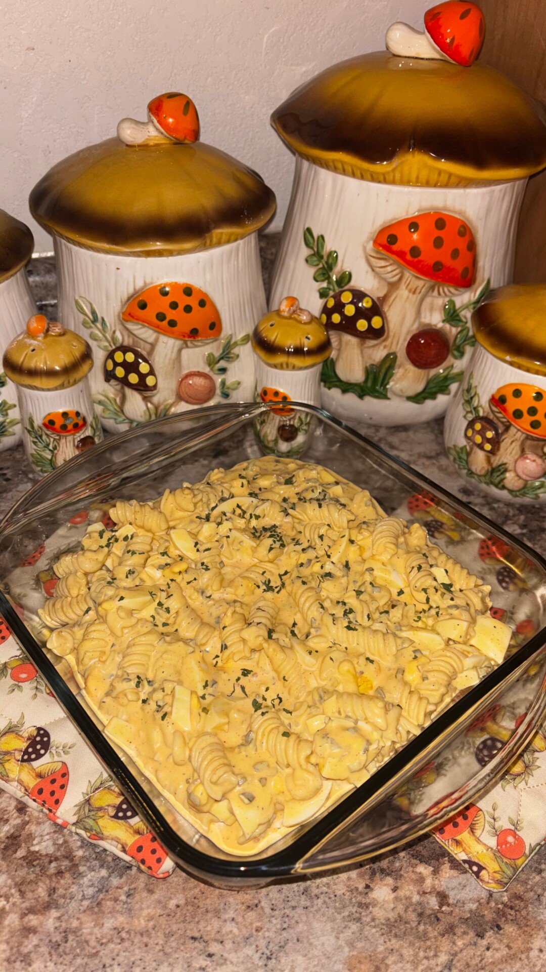 A glass baking dish filled with creamy, yellow rotini pasta sits on a counter, garnished with herbs. Behind it are ceramic mushroom-themed canisters in various sizes, decorated with orange and yellow mushroom tops.