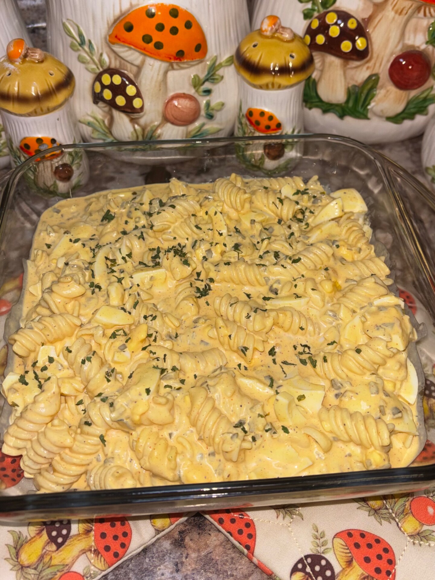 A square glass dish filled with creamy rotini pasta salad, topped with herbs. In the background, there are decorative ceramic mushroom containers and a patterned cloth with mushrooms and strawberries.