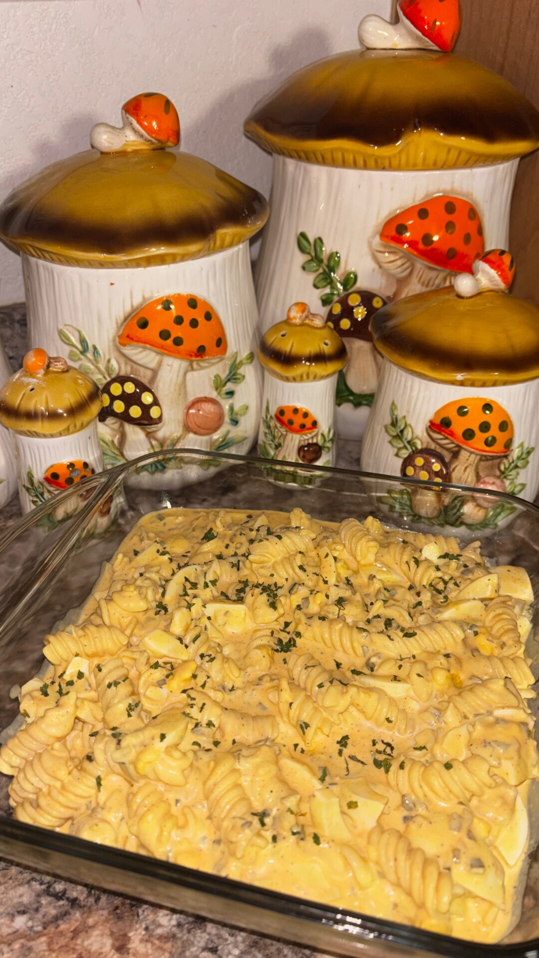A glass baking dish filled with creamy rotini pasta, garnished with herbs, sits on a countertop in front of a set of vintage ceramic canisters decorated with colorful mushrooms.
