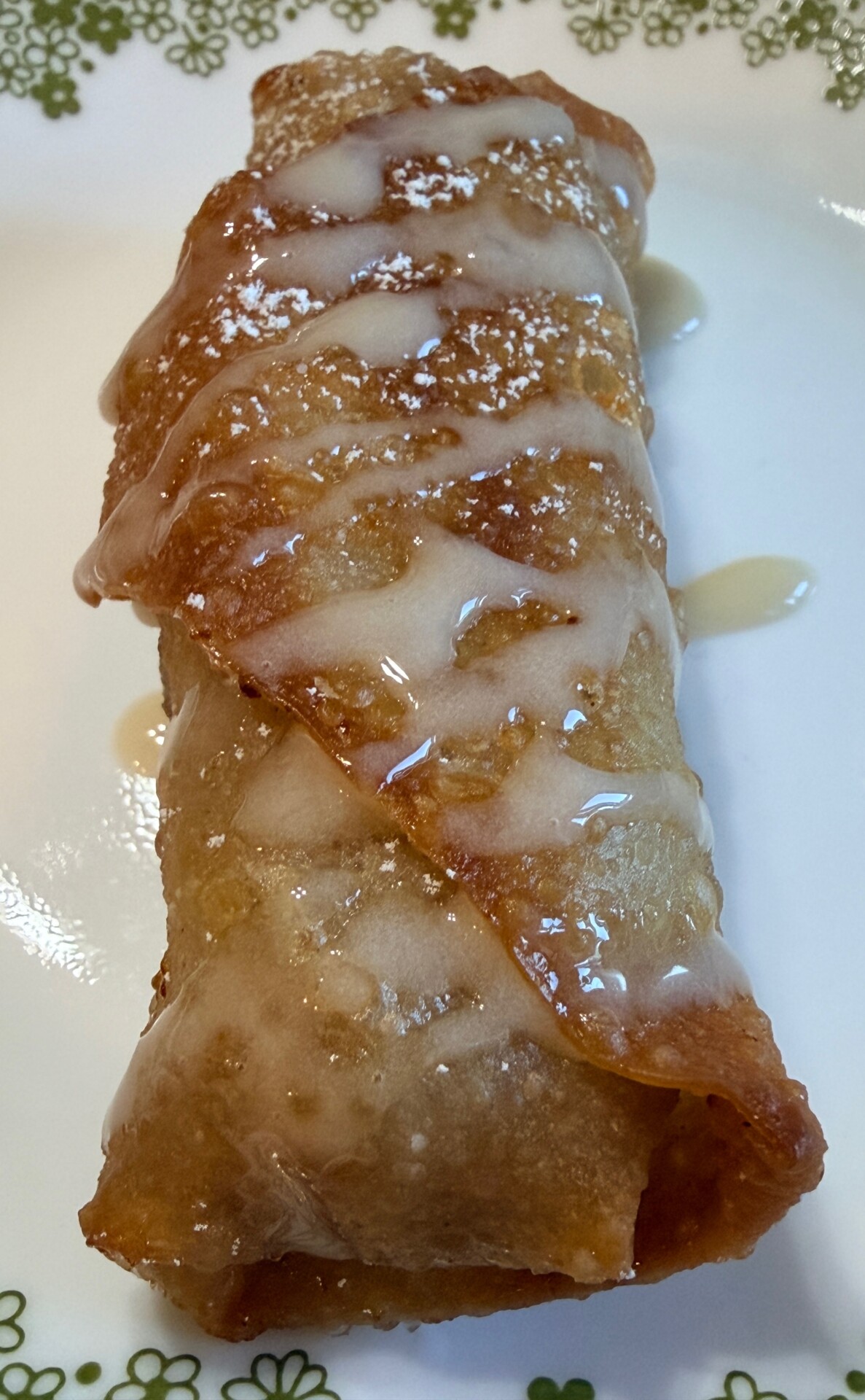 A close-up of a golden-brown, crispy rolled pastry drizzled with white icing on a white plate with a green floral edge pattern.