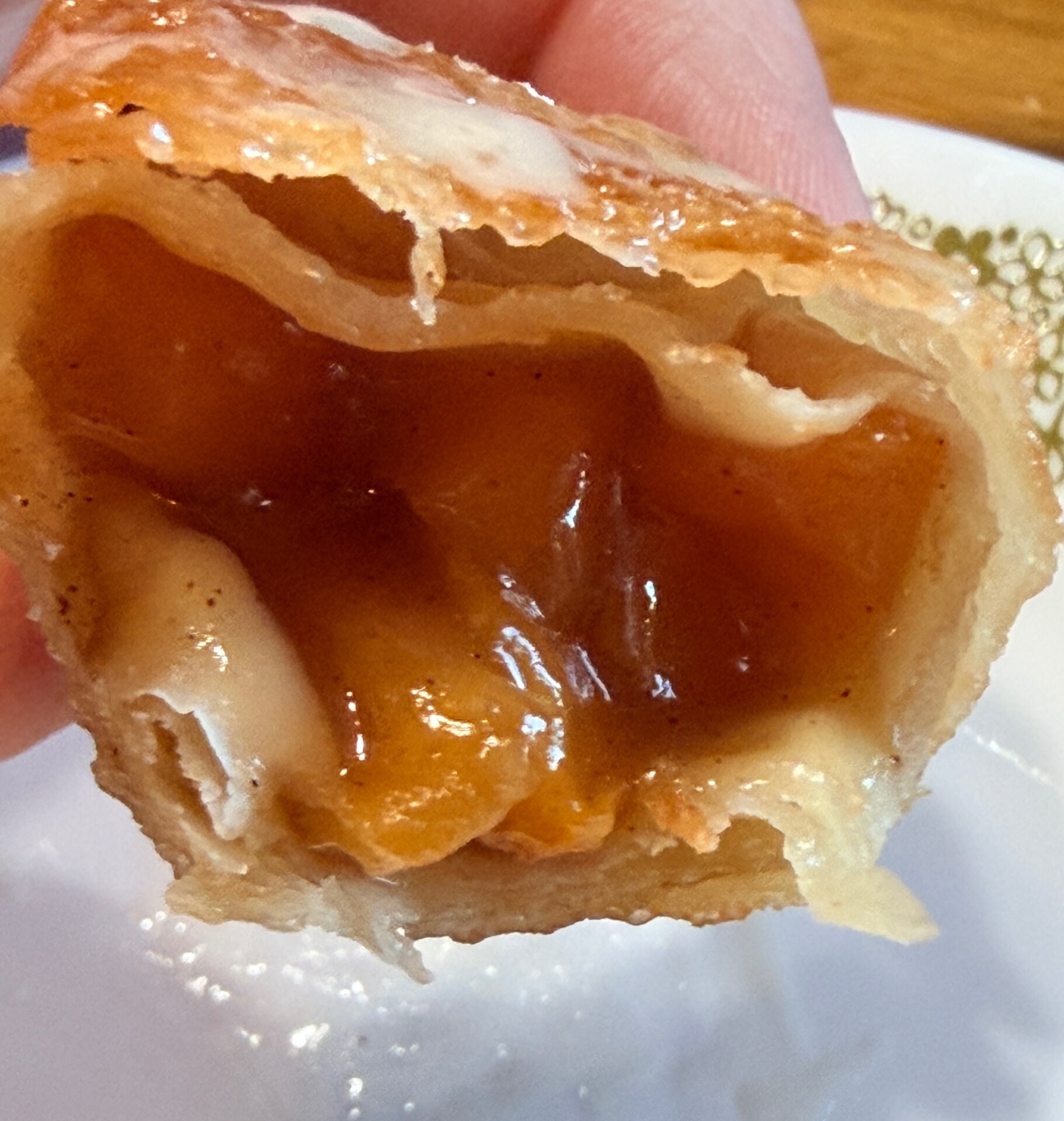 A close-up of a hand holding a fried pastry with a golden, flaky crust, showing a gooey, cinnamon-spiced apple filling inside. The pastry is partially eaten, exposing its juicy interior.