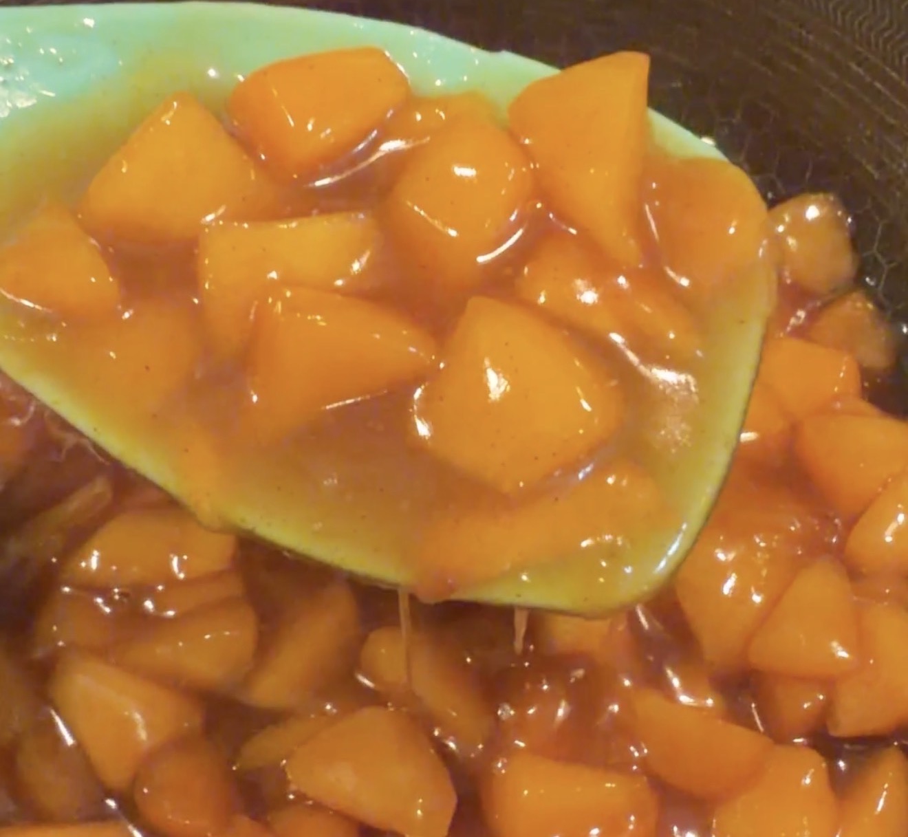 A close-up of a spoon lifting glossy, diced cooked apples coated in a thick, golden-brown sauce, likely being prepared for a dessert.