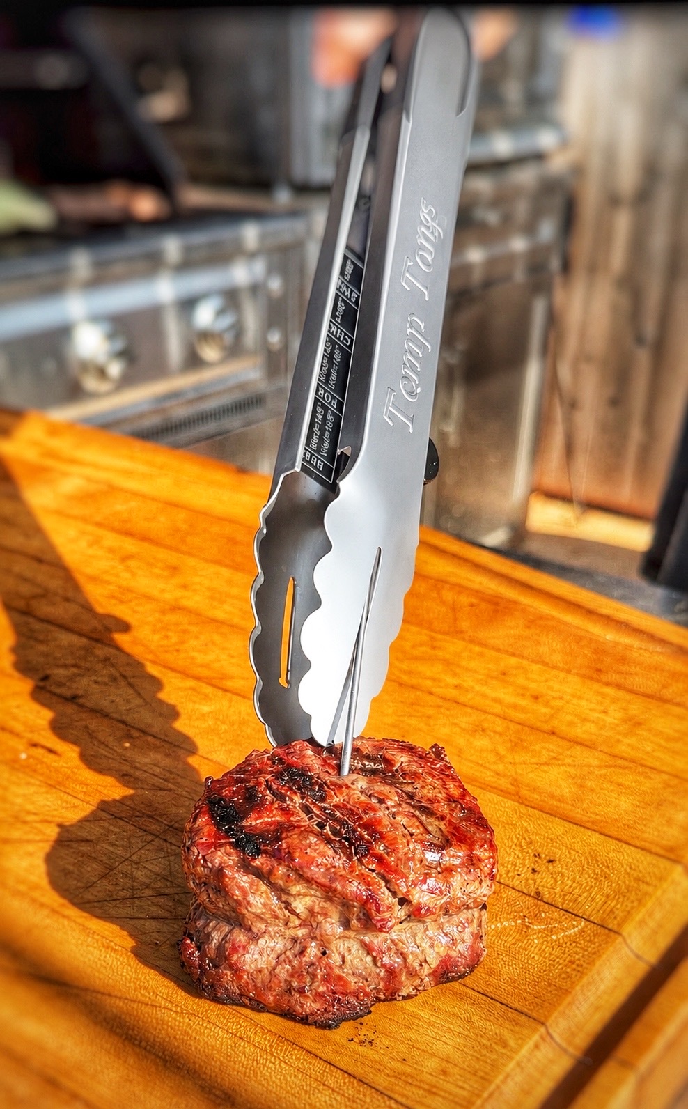 A pair of metal tongs labeled Funny Tongs holds a cooked steak on a wooden cutting board, with a kitchen stove and oven in the background.