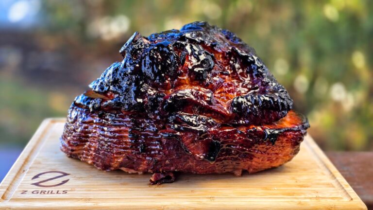 A glazed, spiral-cut ham with a caramelized, slightly charred crust sits on a wooden cutting board outdoors, with a blurred green background.