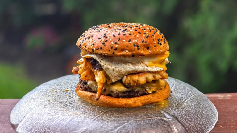 A cheeseburger with a sesame seed bun, topped with a fried egg, crinkle-cut fries, and melted cheese sits on a glass plate outdoors.