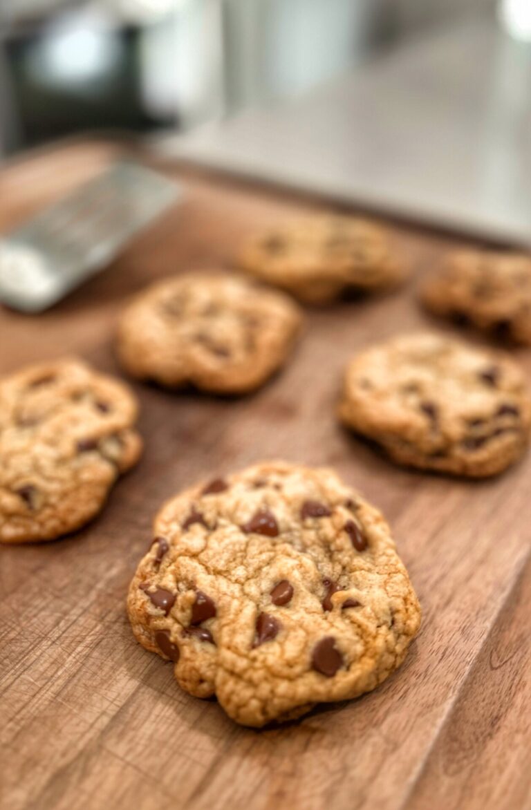 A close-up of several chocolate chip cookies on a wooden surface, with one cookie in sharp focus and the rest softly blurred in the background.