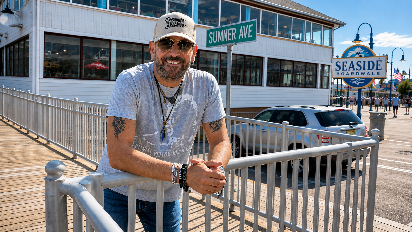 A man in sunglasses and a cap leans on a railing at Seaside Boardwalk near a sign for Sumner Ave, with a building, parked cars, and blue sky in the background.