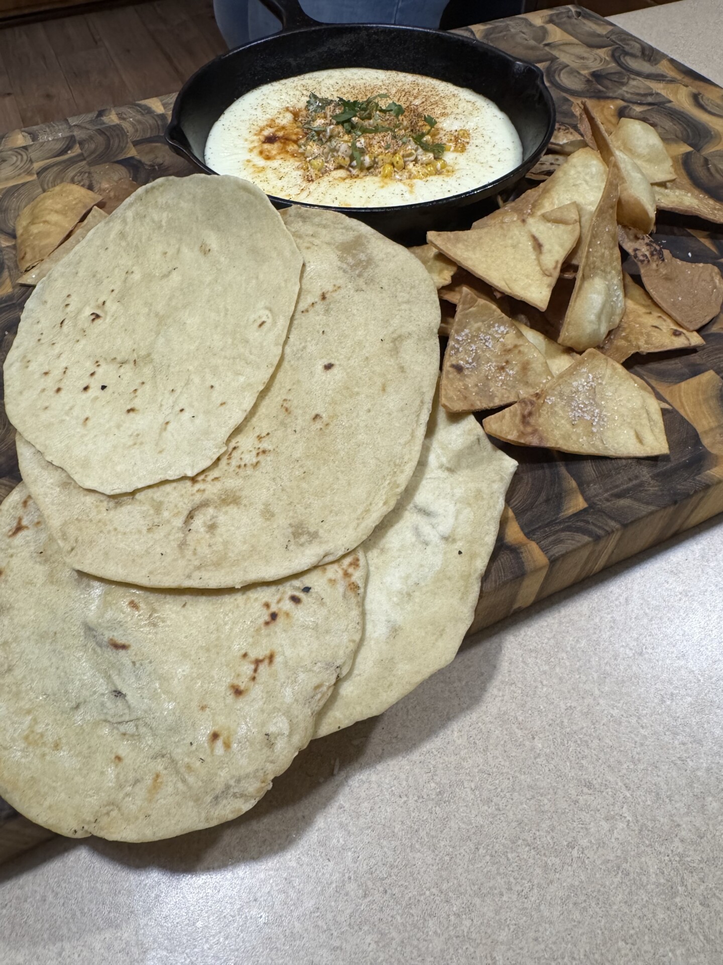 A wooden board with several pieces of flatbread, crispy tortilla chips, and a cast iron skillet filled with creamy dip garnished with herbs and paprika.