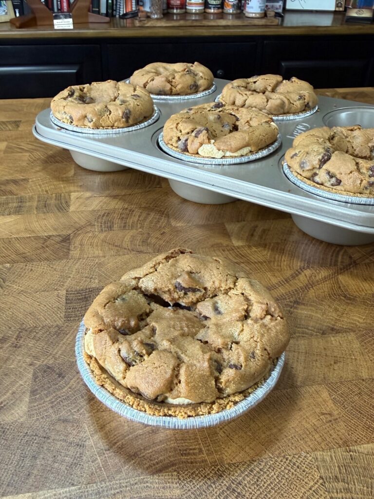 Six round, golden-brown chocolate chip cookie pies sit on a wooden counter. Five are in a muffin tin, while one is placed in front, outside of the tray, closer to the camera.