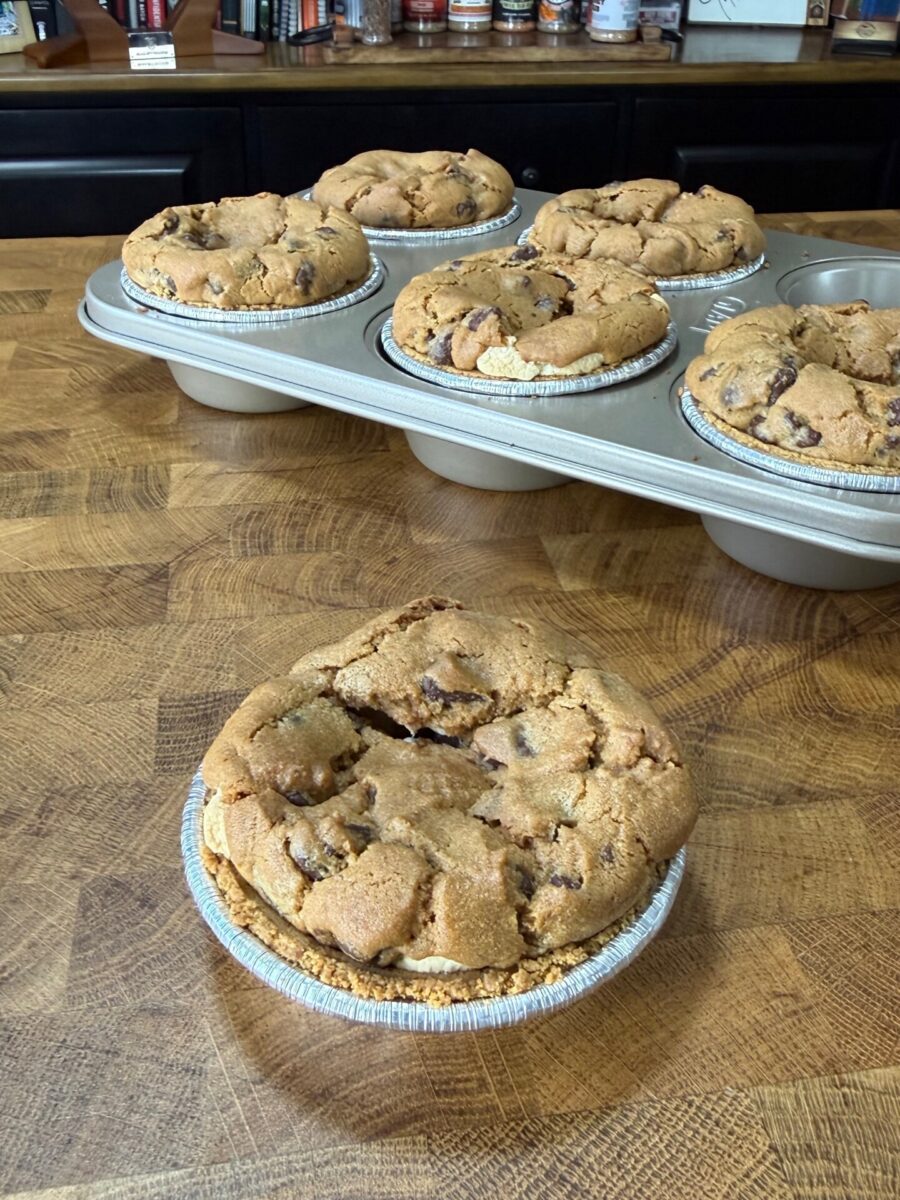 Six round, golden-brown chocolate chip cookie pies sit on a wooden counter. Five are in a muffin tin, while one is placed in front, outside of the tray, closer to the camera.
