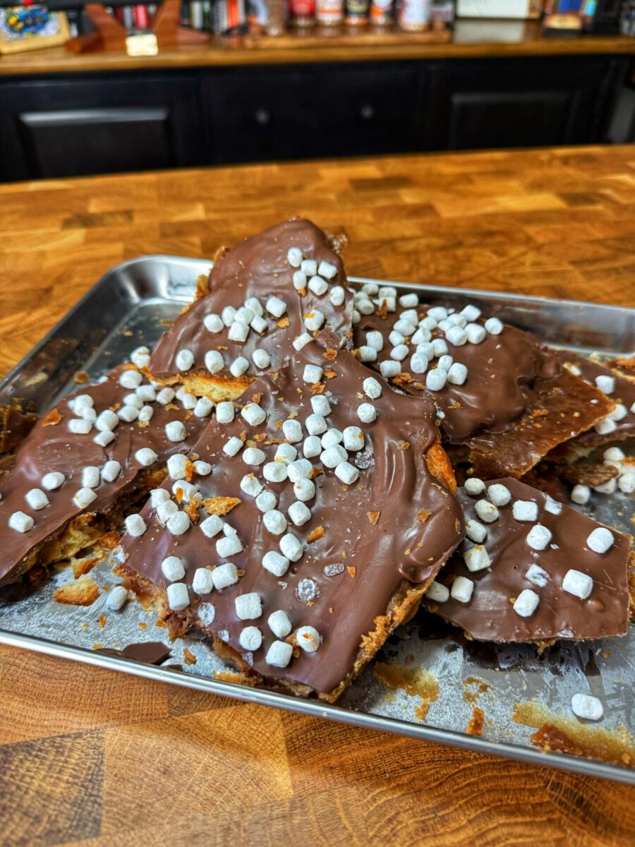 A metal tray holds pieces of chocolate-covered toffee topped with mini marshmallows, displayed on a wooden counter in a kitchen setting.
