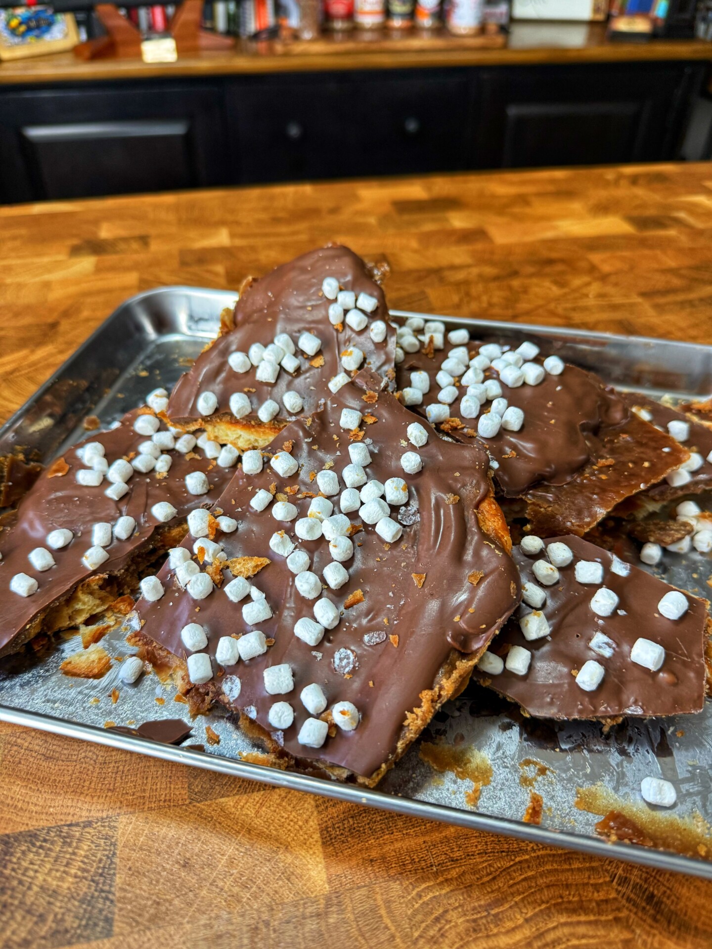 A metal tray holds pieces of chocolate-covered toffee topped with mini marshmallows, displayed on a wooden counter in a kitchen setting.