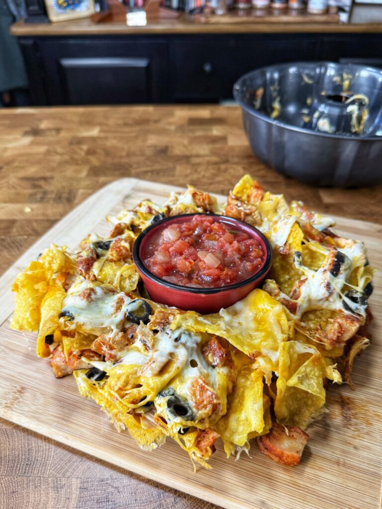A pile of cheesy nachos with grilled chicken and olives is served on a wooden board, topped with a bowl of chunky salsa in the center. A metal mixing bowl and spices are visible in the background.