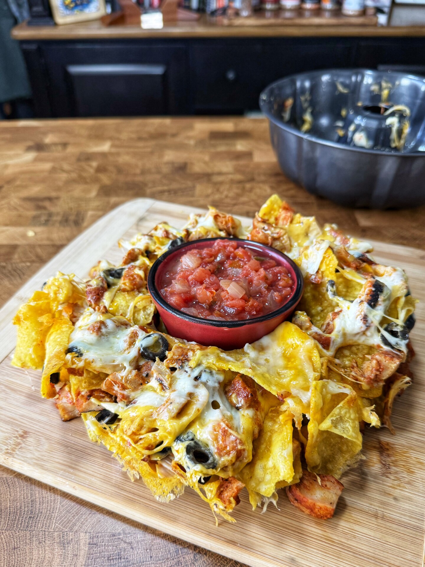 A pile of cheesy nachos with grilled chicken and olives is served on a wooden board, topped with a bowl of chunky salsa in the center. A metal mixing bowl and spices are visible in the background.