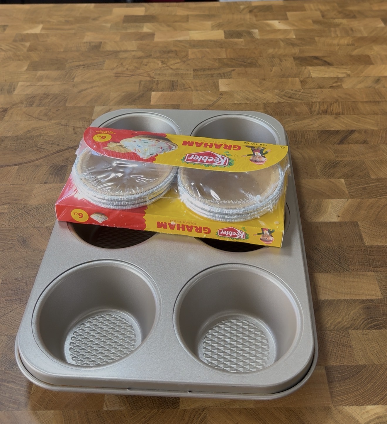 A muffin tin sits on a wooden surface with a pack of six aluminum graham pie crusts in colorful packaging placed on top of it.