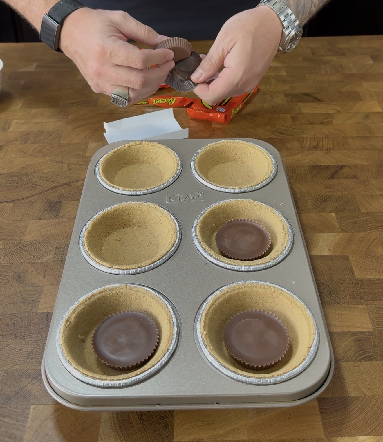 A person places peanut butter cups into graham cracker crusts in a muffin tin, preparing a dessert on a wooden countertop.
