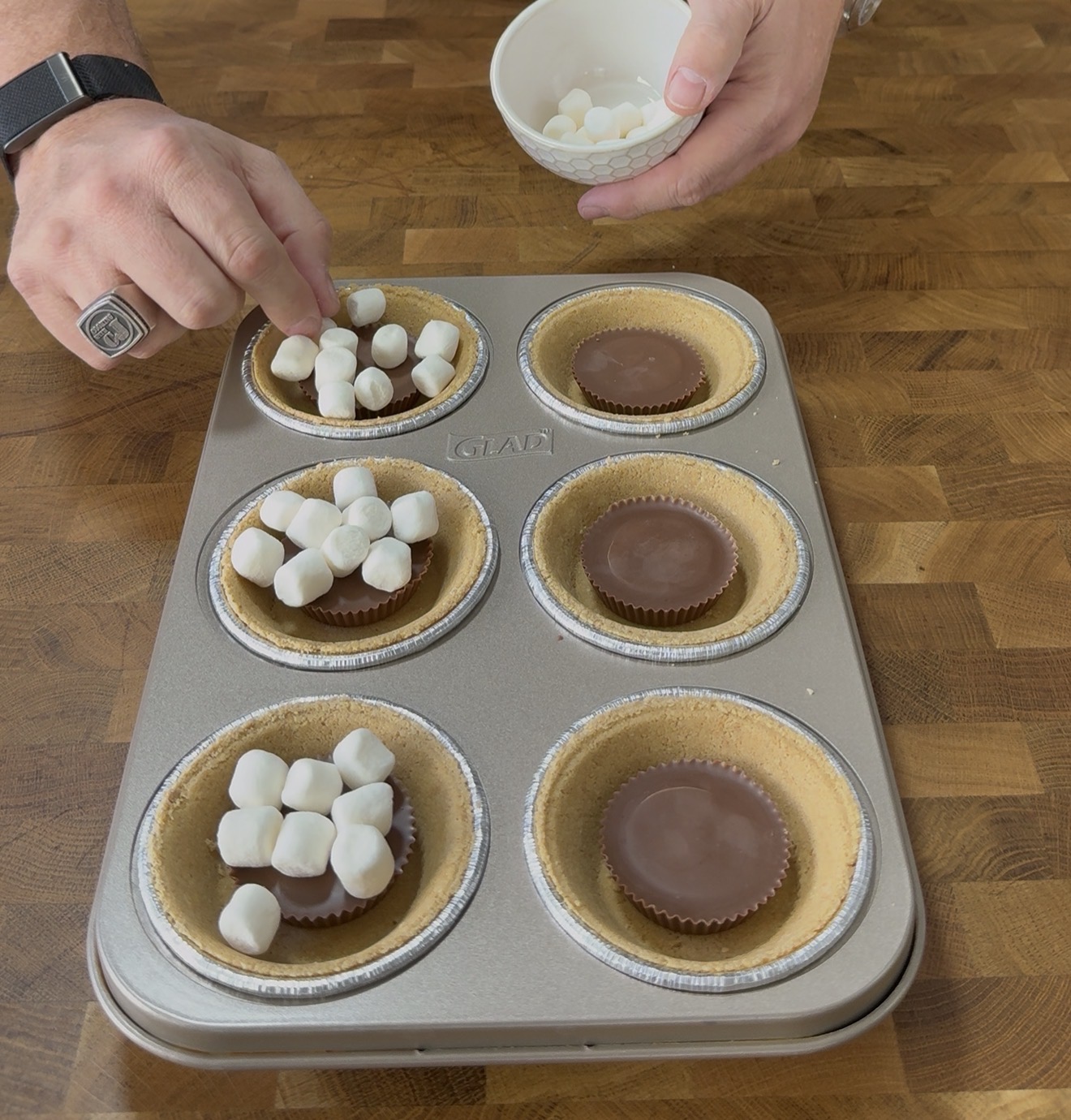 A person places mini marshmallows into graham cracker pie crusts, each with a chocolate peanut butter cup, using a muffin tin on a wooden surface.