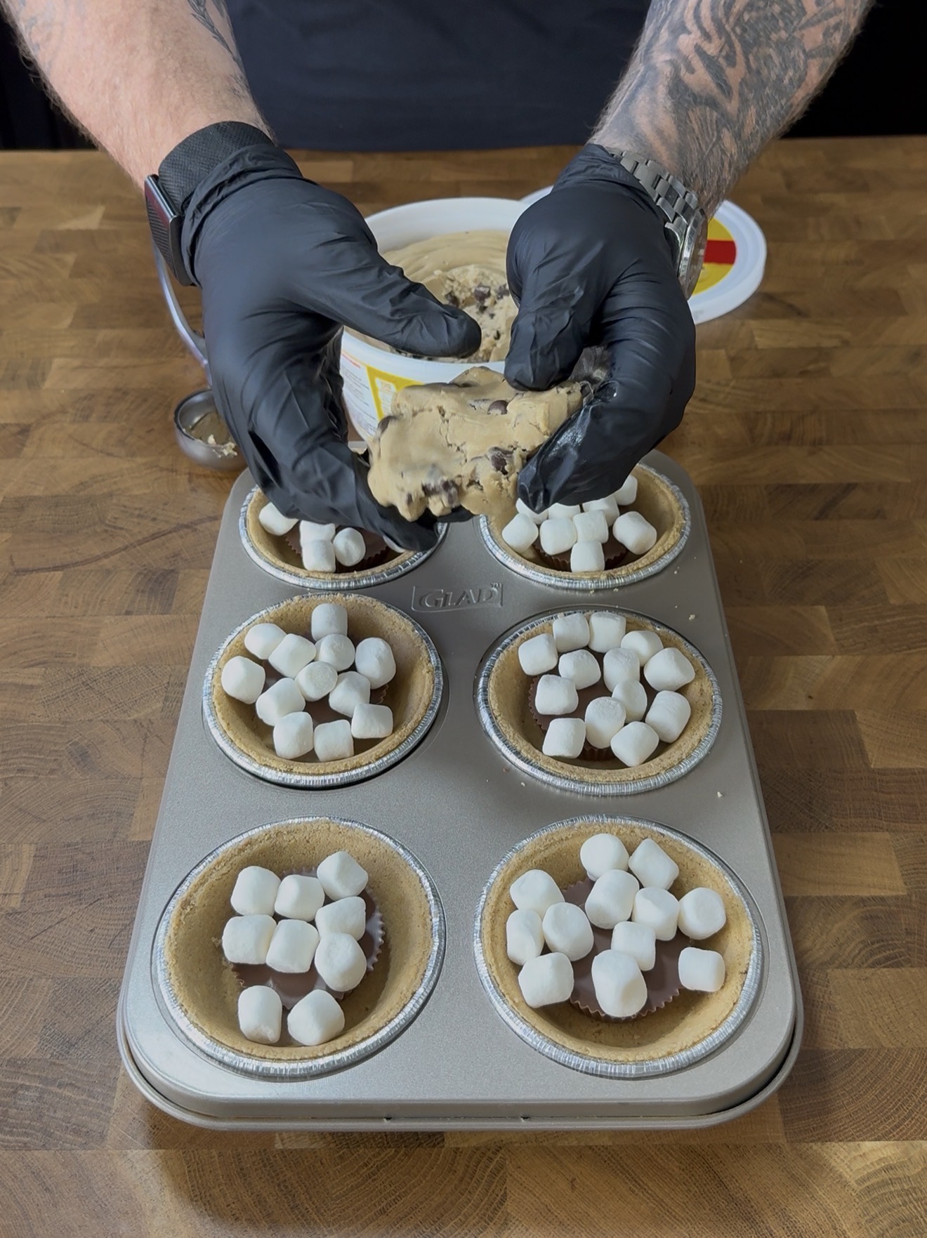A person wearing black gloves presses cookie dough over mini pie crusts filled with marshmallows in a muffin tray, on a wooden surface.