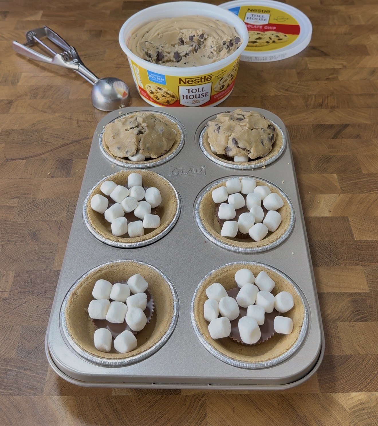 A muffin tin holds six graham crusts, three filled with marshmallows and three topped with baked cookie dough. A tub of Nestlé Toll House cookie dough and a scoop sit in the background on a wooden surface.