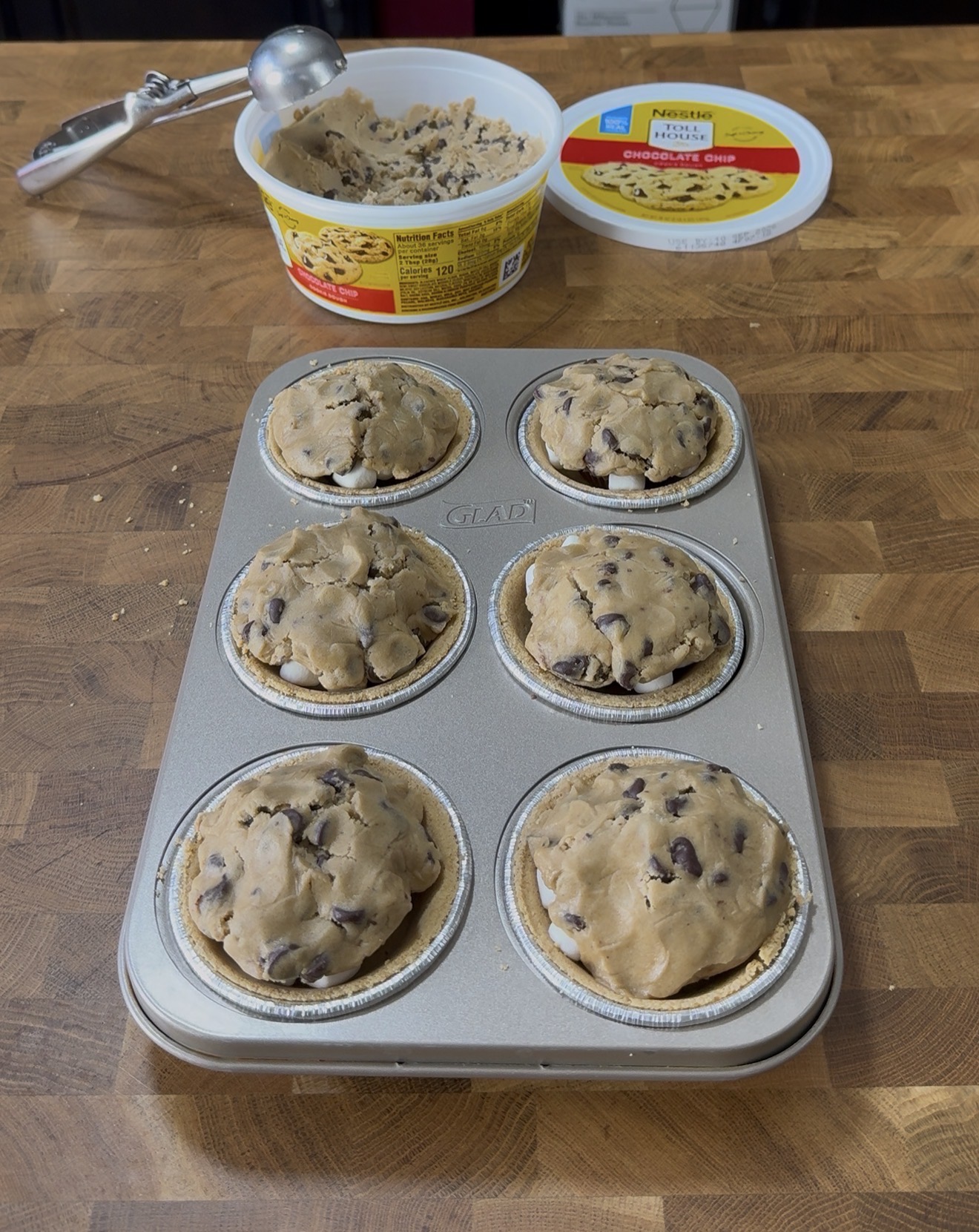 A muffin tin with six wells filled with cookie dough sits on a wooden countertop. An open tub of Nestlé Toll House cookie dough and an ice cream scoop are in the background.
