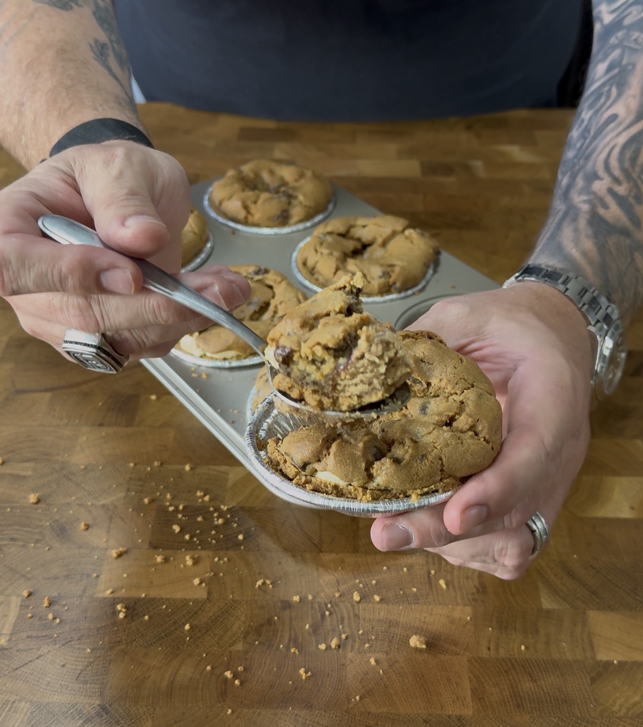 A person with tattoos holds a spoonful of cookie pie above a muffin tin filled with more cookie pies on a wooden table. Crumbs are scattered on the table.