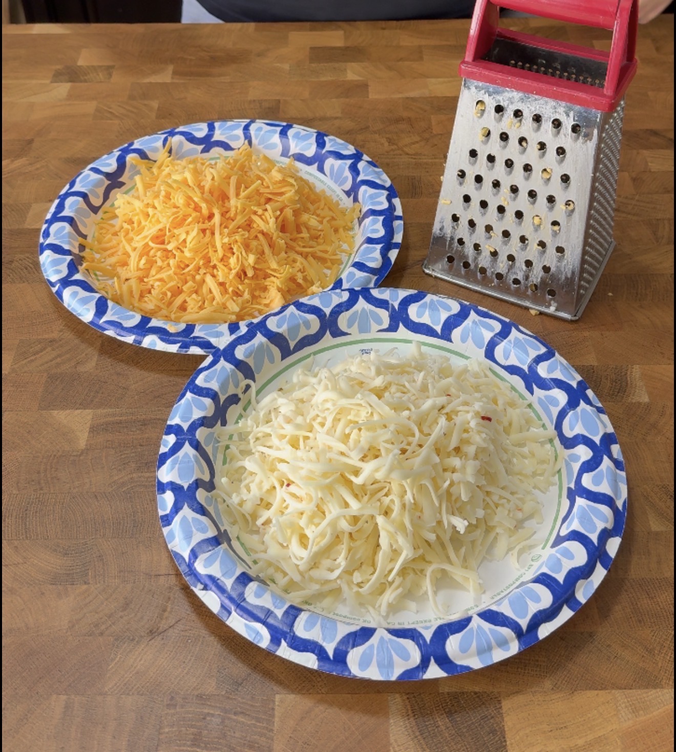 Two paper plates on a wooden surface hold shredded yellow and white cheese. A metal box grater with a red handle is next to the plates.