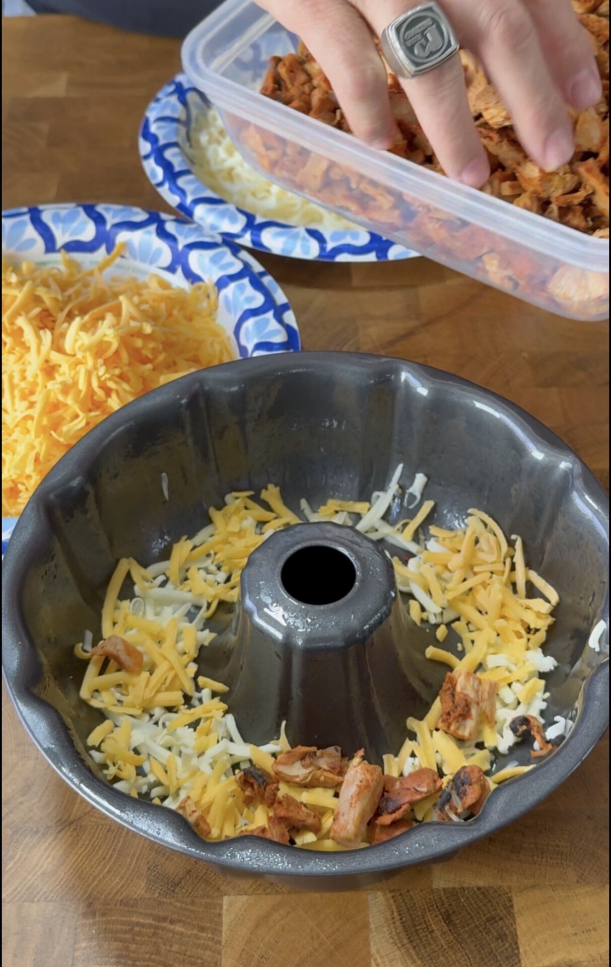 A hand adds shredded meat from a container into a Bundt pan, which already contains cheese and meat. Next to the pan are plates with shredded cheese and more meat.