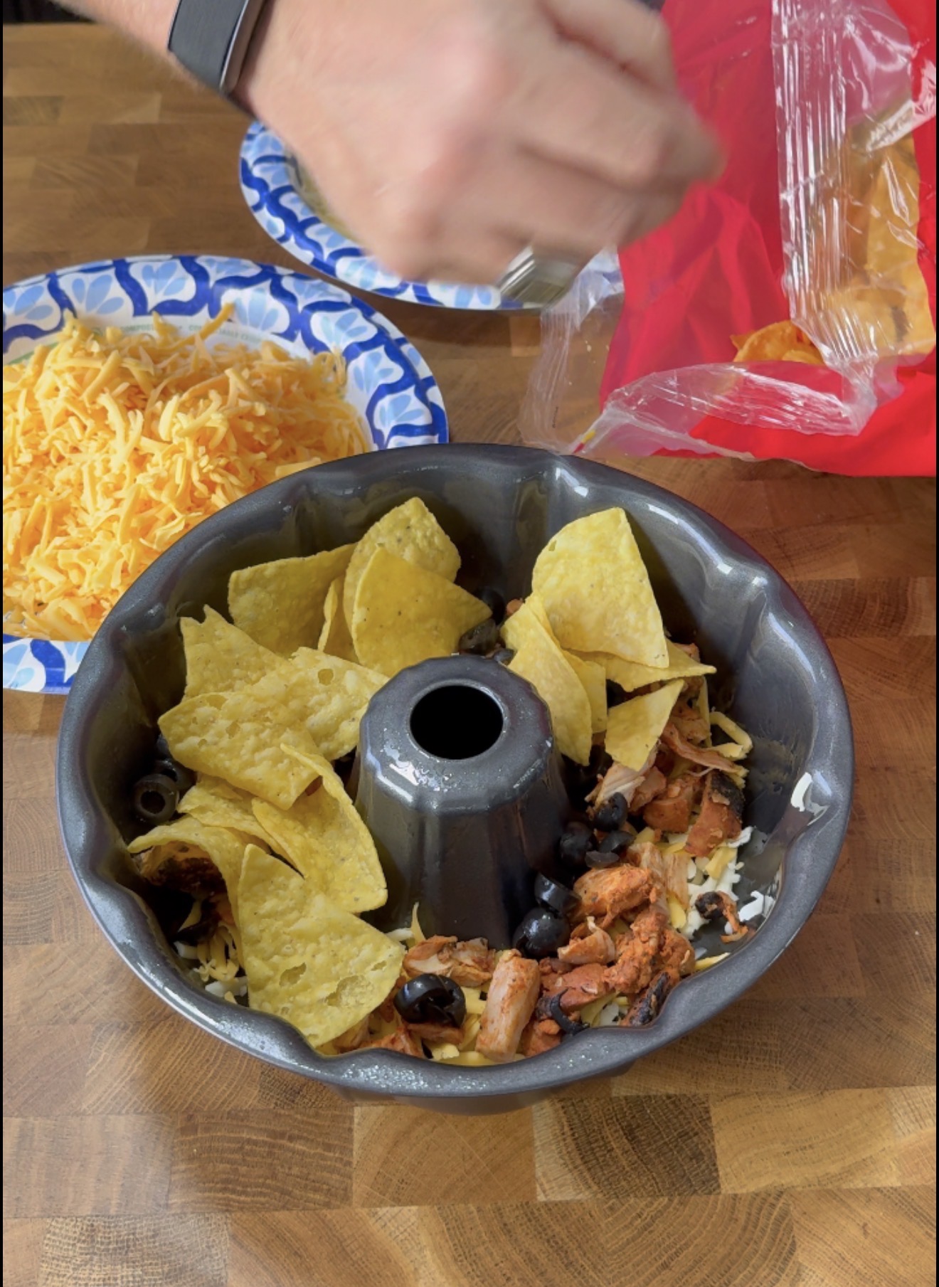 A bundt pan with tortilla chips, chicken, black olives, and cheese inside. A hand with a watch holds chips, with shredded cheese and chip bag on the wooden counter nearby.