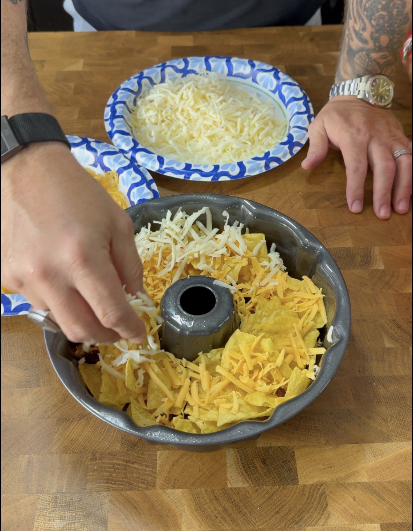 A person sprinkles shredded cheese over tortilla chips in a bundt pan, with more shredded cheese on a paper plate nearby, on a wooden surface.